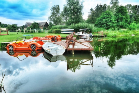 Sommerhaus am See mit Tretbooten, Ruderboot und Steg, umgeben von grüner Natur und ruhigem Wasser.