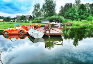 Sommerhaus am See mit Tretbooten, Ruderboot und Steg, umgeben von grüner Natur und ruhigem Wasser.
