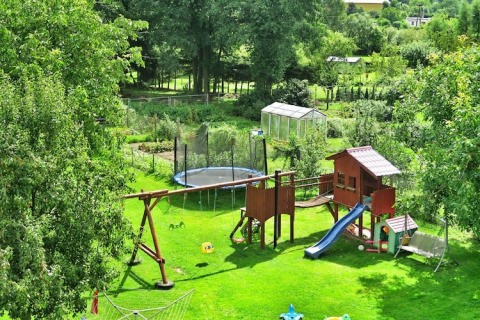 Spielplatz mit Schaukeln, Rutsche und Trampolin in einem grünen Garten mit Gewächshaus im Hintergrund.