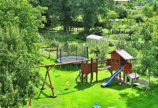 Spielplatz mit Schaukeln, Rutsche und Trampolin in einem grünen Garten mit Gewächshaus im Hintergrund.