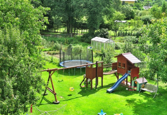 Legeplads med gynger, rutsjebane og trampolin i en grøn have tæt på et drivhus og træer.