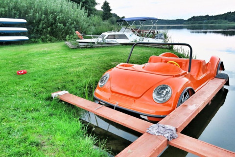 Ein orangefarbenes tretboot in Auto-Optik liegt am Ufer eines Sees mit Booten im Hintergrund.
