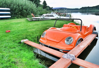 Ein orangefarbenes tretboot in Auto-Optik liegt am Ufer eines Sees mit Booten im Hintergrund.