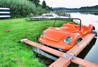 An orange pedal boat shaped like a car is docked at the lakeside with other boats and nature around.