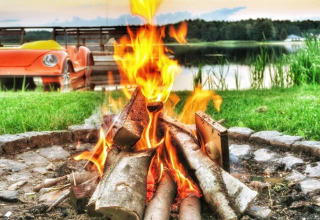 Lagerfeuer brennt vor einem See mit orangefarbenem Auto im Hintergrund, Domek letniskowy PATRYK Ostrowiec Slawienski.
