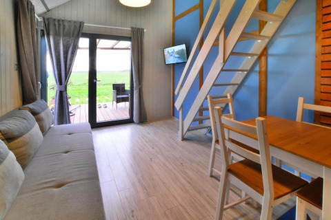 Indvendigt billede af en hyggelig lodge med træmøbler, sofa, trappe til hems og udsigt mod naturen.