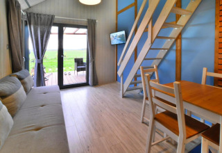 Indvendigt billede af en hyggelig lodge med træmøbler, sofa, trappe til hems og udsigt mod naturen.