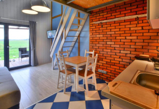 Hyggelig lodge med murstensvæg, spisebord, køkken og trappe til hems samt udsigt til det fri