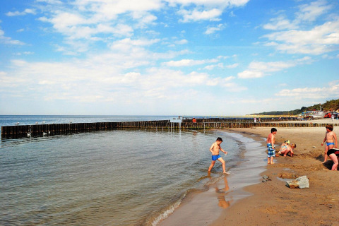 Kinderen spelen op het strand en in ondiep water bij houten golfbrekers, onder een blauwe lucht.