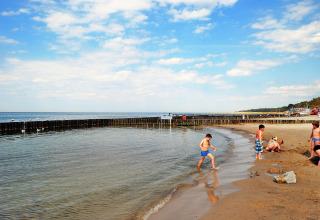 Kinderen spelen op het strand en in ondiep water bij houten golfbrekers, onder een blauwe lucht.