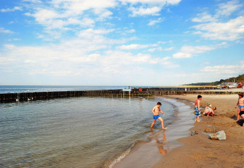 Niños jugando en la playa arenosa y en el agua, junto a espigones de madera, bajo cielo azul.