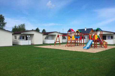 Vrolijk speelplein met glijbanen en vakantiehuisjes rondom, gelegen op een groene grasmat, blauwe lucht.
