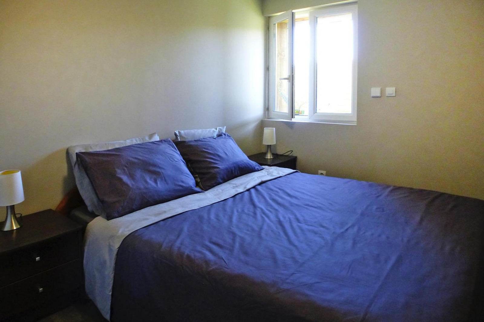 Slaapkamer met tweepersoonsbed, donkerblauw beddengoed, nachtkastjes en open raam in Holiday home «Le Porge», Frankrijk.