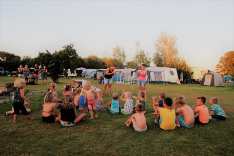 Kinderen zitten en spelen op het gras bij Minicamping Falkenborg met caravans en volwassenen op de achtergrond.