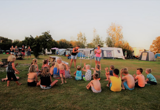 Kinderen zitten en spelen op het gras bij Minicamping Falkenborg met caravans en volwassenen op de achtergrond.