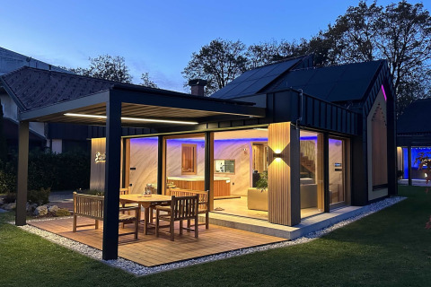 Modern Slovenian lodge with illuminated patio, outdoor dining set, large windows and cozy interior at dusk.