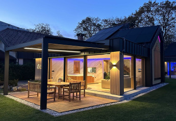 Moderna cabaña en Eslovenia con terraza iluminada, comedor exterior y grandes ventanales al anochecer.