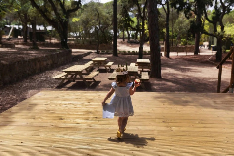 Petite fille couronnée avec une pomme marchant vers des tables de pique-nique à Wecamp Santa Cristina, Catalogne.