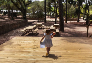 Young girl with a crown and apple walks towards picnic tables at the forested Wecamp Santa Cristina in Catalonia.