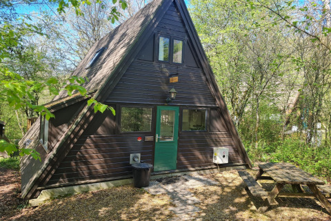 Dreieckige Berghütte mit grüner Tür im Wald, Picknickbank und Beschilderung Chalet no 142 im Frühling.