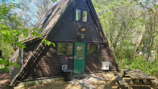 Dreieckige Berghütte mit grüner Tür im Wald, Picknickbank und Beschilderung Chalet no 142 im Frühling.