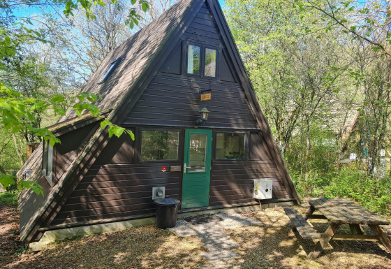 Dreieckige Berghütte mit grüner Tür im Wald, Picknickbank und Beschilderung Chalet no 142 im Frühling.