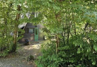 Fassade von Chalet Nr. 142 in Belgien, teils verborgen zwischen dichten, grünen Bäumen und Laub.