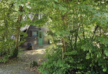 Fassade von Chalet Nr. 142 in Belgien, teils verborgen zwischen dichten, grünen Bäumen und Laub.