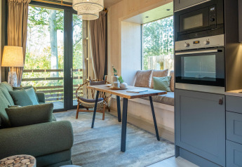 Interno di tiny house moderna con divano verde, tavolo da pranzo, finestrone e cucina compatta.