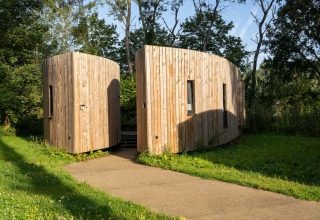 Et moderne glamping-ophold i naturen med to halvrunde træhytter omgivet af grønne træer og græs.