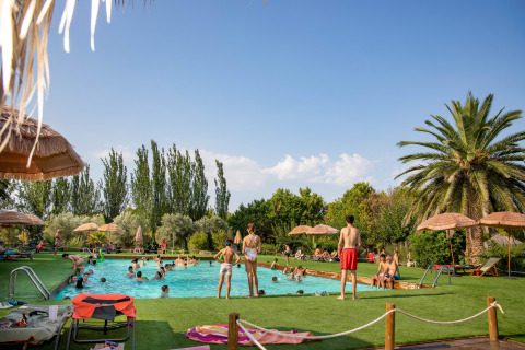 Parque vacacional en Aragón, España, con piscina, palmeras y visitantes disfrutando en TAIGA Lake Caspe al sol.