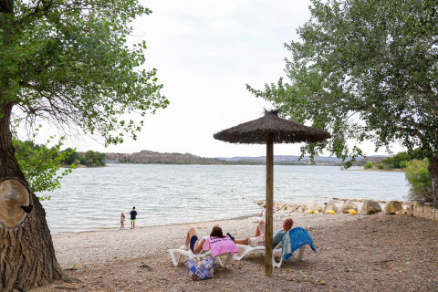 People lounging under a thatched parasol by the shore at TAIGA Lake Caspe holiday park in Aragón, Spain.