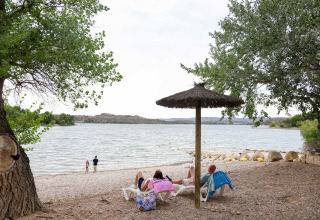 People lounging under a thatched parasol by the shore at TAIGA Lake Caspe holiday park in Aragón, Spain.