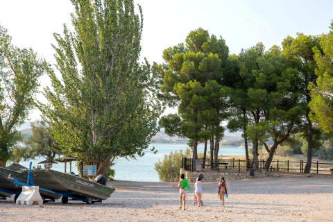Des enfants marchent vers le lac au parc de vacances TAIGA Lake Caspe, en Aragón, Espagne, entourés d’arbres et de bateaux.