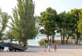 Des enfants marchent vers le lac au parc de vacances TAIGA Lake Caspe, en Aragón, Espagne, entourés d’arbres et de bateaux.