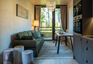 Accogliente tiny house moderna con divano verde, tavolo da pranzo e grande finestra vista bosco.