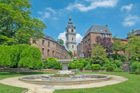 Vista di un parco verde con fontana e edifici storici sotto un cielo azzurro in Belgio d'estate.
