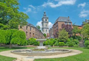 Vista di un parco verde con fontana e edifici storici sotto un cielo azzurro in Belgio d'estate.