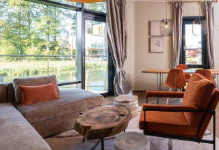 Moderne lodge-stue med store vinduer, udsigt til skov og flod, rustikke træborde, orange stol og spiseplads.