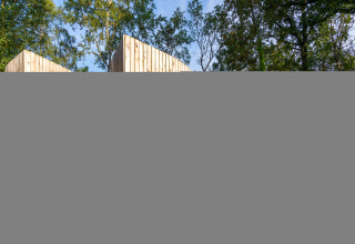 Cabaña de madera 'The Lodge by YourNature' en Bélgica, rodeada de árboles y cielo azul.