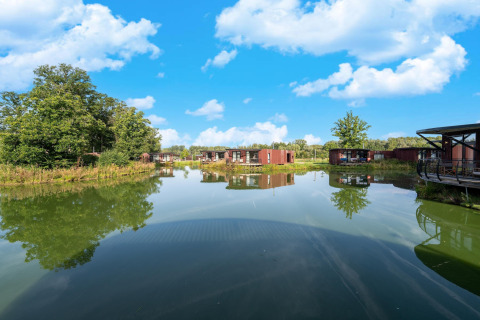 Vista de lago tranquilo con modernas cabañas glamping y vegetación bajo un cielo azul y soleado.