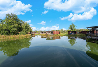 Vista de lago tranquilo con modernas cabañas glamping y vegetación bajo un cielo azul y soleado.