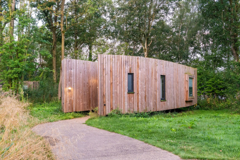 Træbeklædt tiny house, The Lodge af YourNature, omgivet af grønne træer og græs i naturligt miljø.