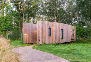 Træbeklædt tiny house, The Lodge af YourNature, omgivet af grønne træer og græs i naturligt miljø.