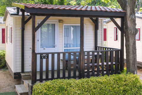 Mobilhome ved Camping Solmar, COTTAGE B, med træveranda, udendørs siddeplads og grøn hæk foran.