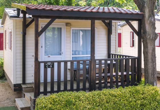 Mobilhome ved Camping Solmar, COTTAGE B, med træveranda, udendørs siddeplads og grøn hæk foran.