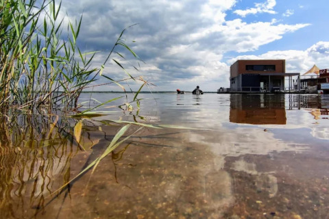 Foto a ras del agua con juncos y una casa flotante en el Gräbendorfer See, Alemania, bajo cielo nublado.