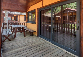 Træveranda med bænk og bord på Camping Solmar - XALET i Spanien, omgivet af træbeklædte hytter.