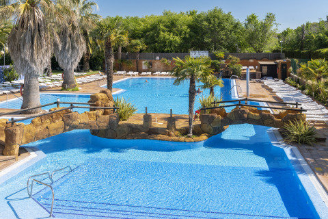 Stort swimmingpoolområde med palmer, stenbro og liggestole på et glampingsted under blå himmel.
