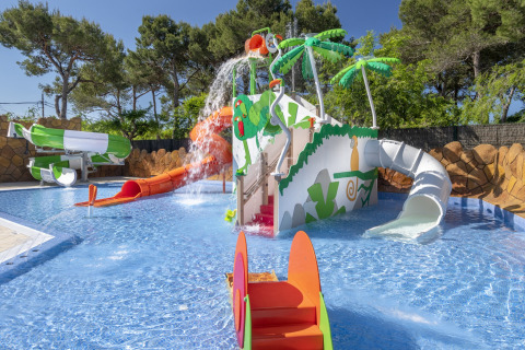 Parco giochi acquatico per bambini con scivoli, palme e spruzzi vicino a una casa mobile sotto il sole.